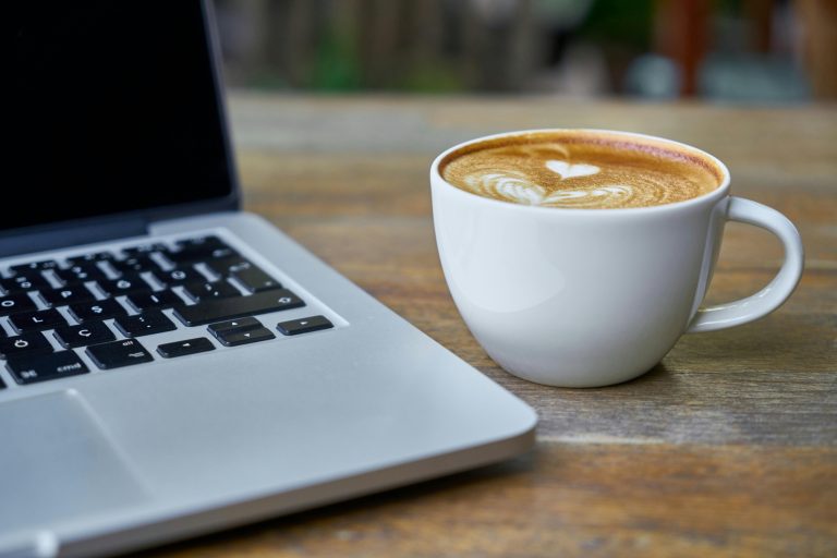 Close-up of a latte coffee with heart art next to a laptop on a rustic wooden table.