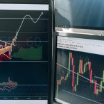 Person pointing at stock market graphs on dual monitors in a modern workspace.