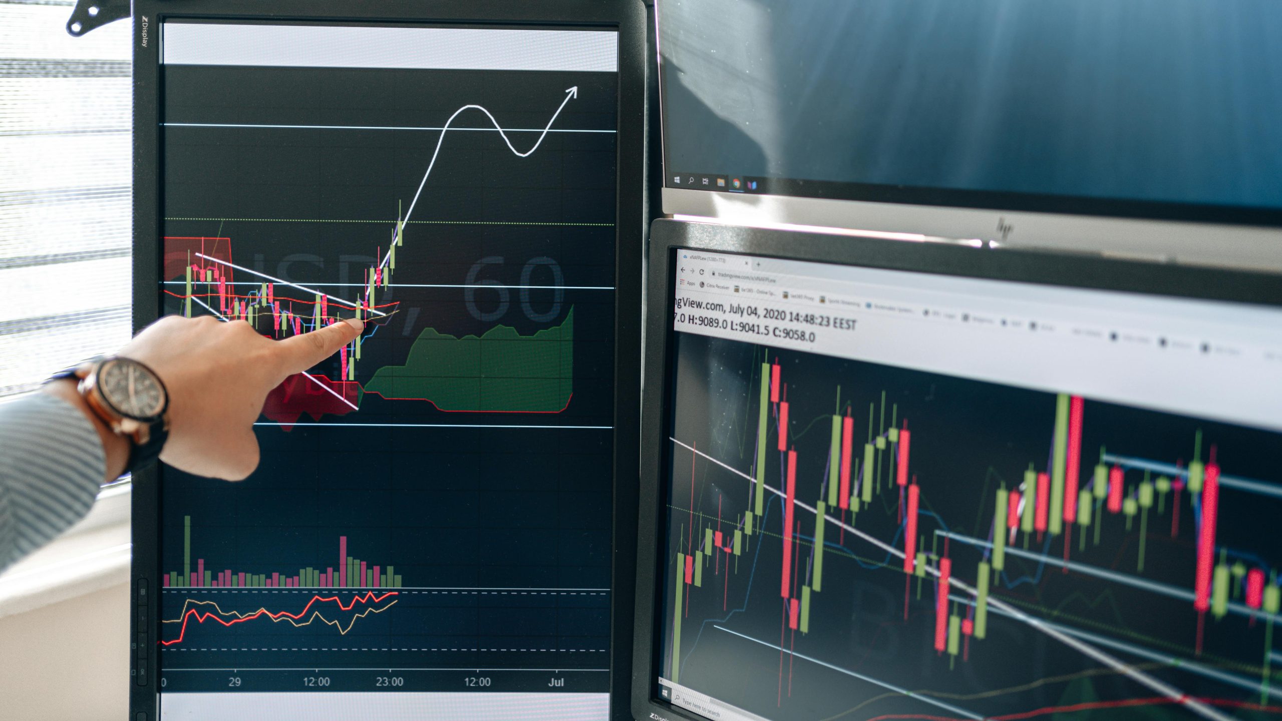 Person pointing at stock market graphs on dual monitors in a modern workspace.